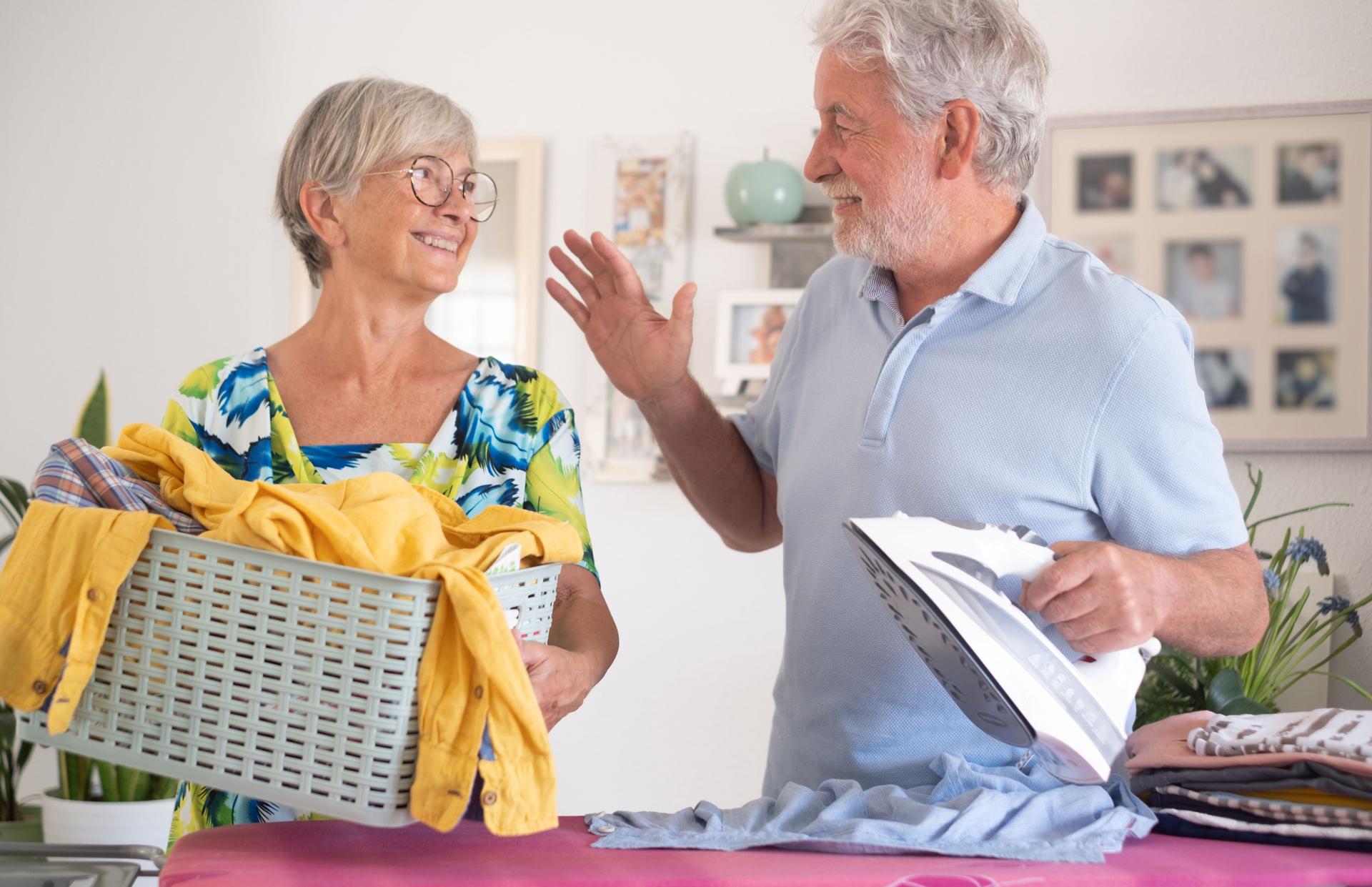 Senior bearded man while ironing at home on the ironing board while his wife bring him new dresses to iron, caucasian happy couple in retirement